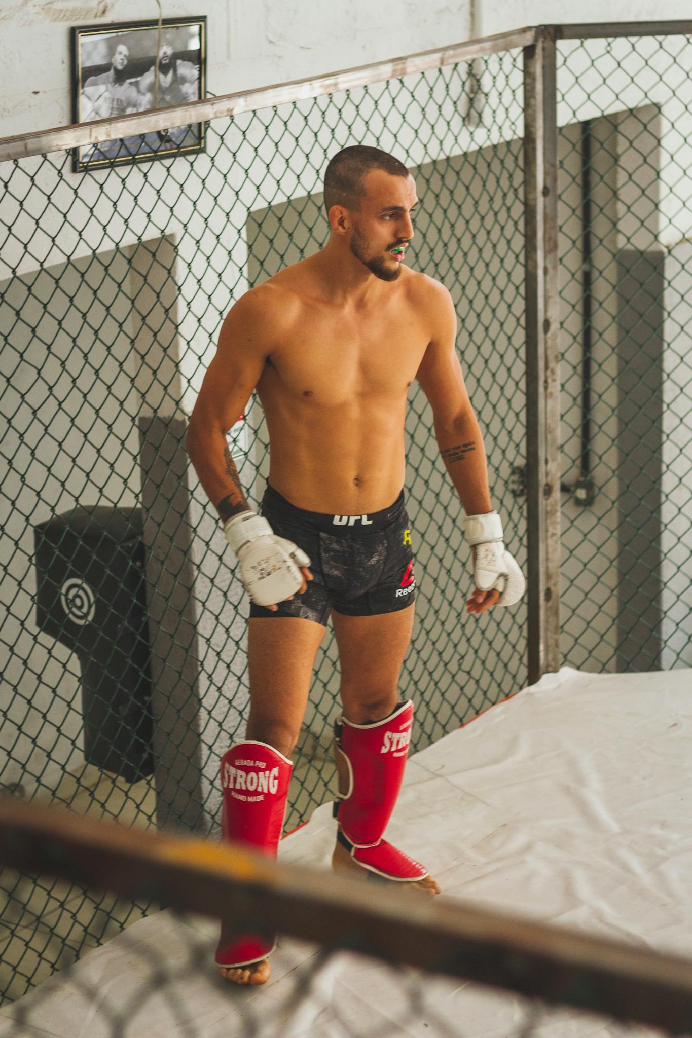 MMA fighter wearing shin guards and gloves preparing for practice in a training cage.