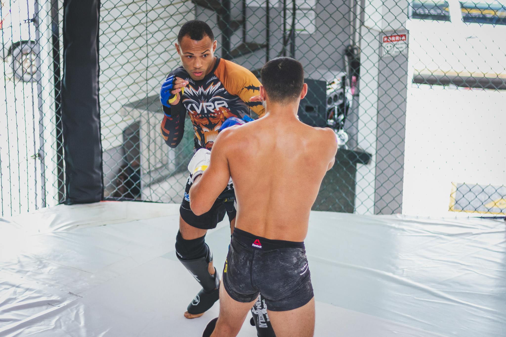 Two athletes engaged in a mixed martial arts sparring session inside a cage.